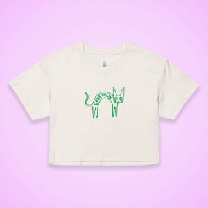 May include: Cream-colored crop top with a green outline drawing of a cat. The word "CAVETOWN" is arched across the cat's back. The shirt has short sleeves and a crew neck. The background is a light purple.