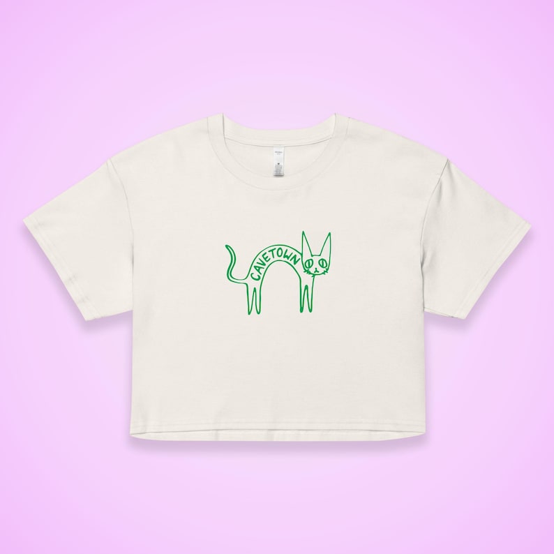 May include: Cream-colored crop top with a green outline drawing of a cat. The word "CAVETOWN" is arched across the cat's back. The shirt has short sleeves and a crew neck. The background is a light purple.