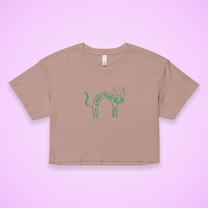May include: A cropped, tan t-shirt featuring a green cartoon cat design. The cat's body spells out "CAVETOWN" with a peace sign in the middle. The shirt has short sleeves and a crew neck, perfect for casual wear.