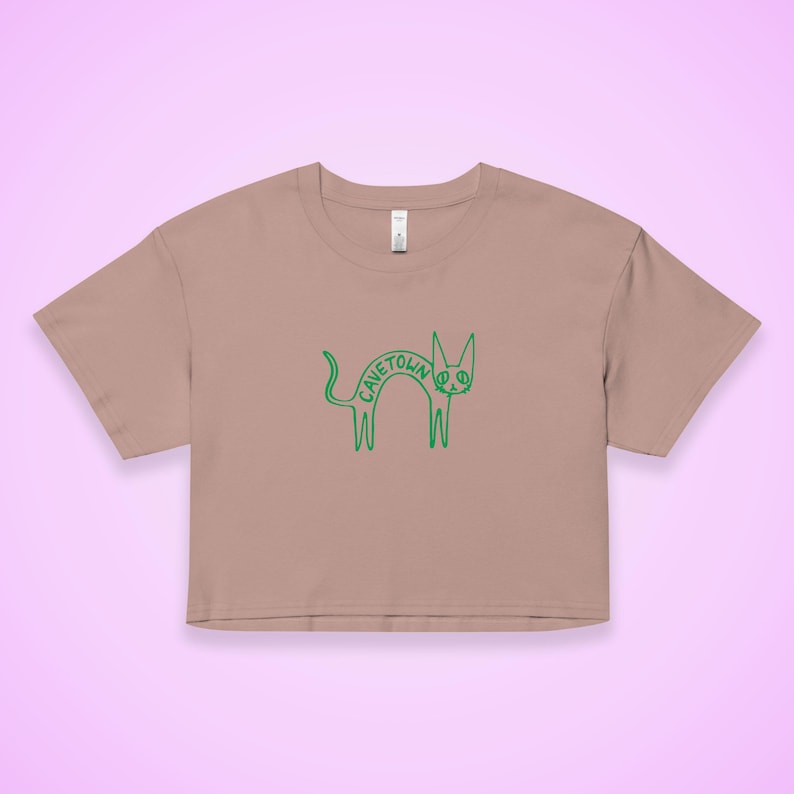 May include: A cropped, tan t-shirt featuring a green cartoon cat design. The cat's body spells out "CAVETOWN" with a peace sign in the middle. The shirt has short sleeves and a crew neck, perfect for casual wear.