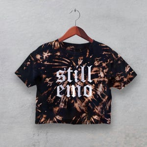 May include: Black and brown tie-dye crop top with the words "still emo" in white gothic lettering. The shirt is hanging on a wooden hanger against a gray background. The shirt has a round neckline and short sleeves.
