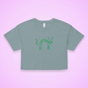 May include: A cropped, light blue t-shirt featuring a green cartoon cat design. The cat has the word "CAVETOWN" written on its body. The design is simple and whimsical, with a peace sign for an eye.