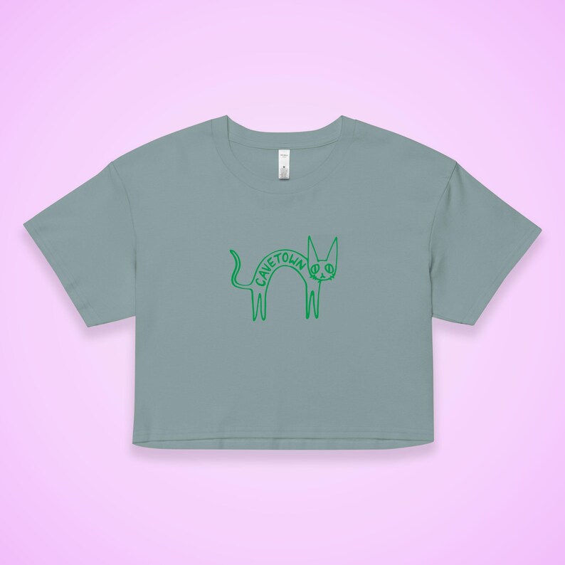 May include: A cropped, light blue t-shirt featuring a green cartoon cat design. The cat has the word "CAVETOWN" written on its body. The design is simple and whimsical, with a peace sign for an eye.
