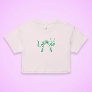 May include: A light pink crop top with a green outline of a cat. The word "CAVETOWN" is written in green across the cat's body. The shirt has short sleeves and a crew neck.