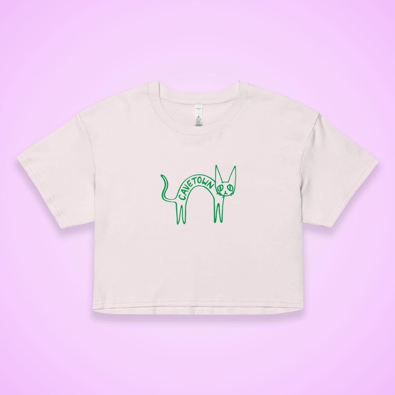 May include: A light pink crop top with a green outline of a cat. The word "CAVETOWN" is written in green across the cat's body. The shirt has short sleeves and a crew neck.