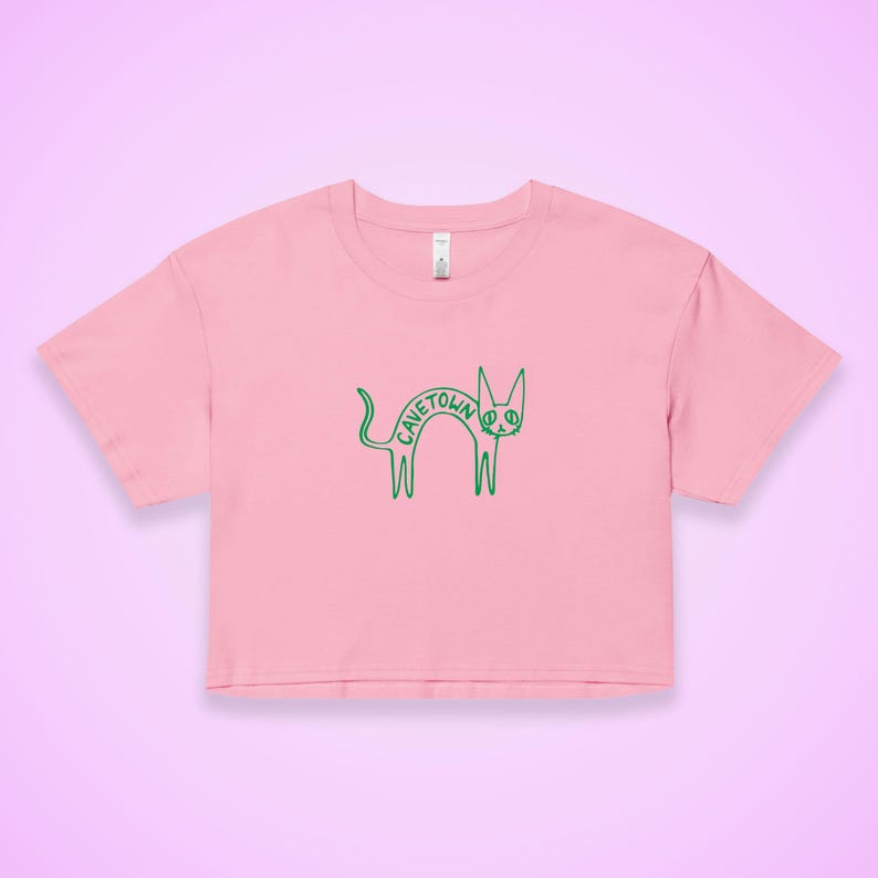 May include: A pink crop top featuring a green cartoon cat design with the word "CAVETOWN" in the center. The cat has a peace sign for an eye. The shirt has short sleeves and a crew neck.