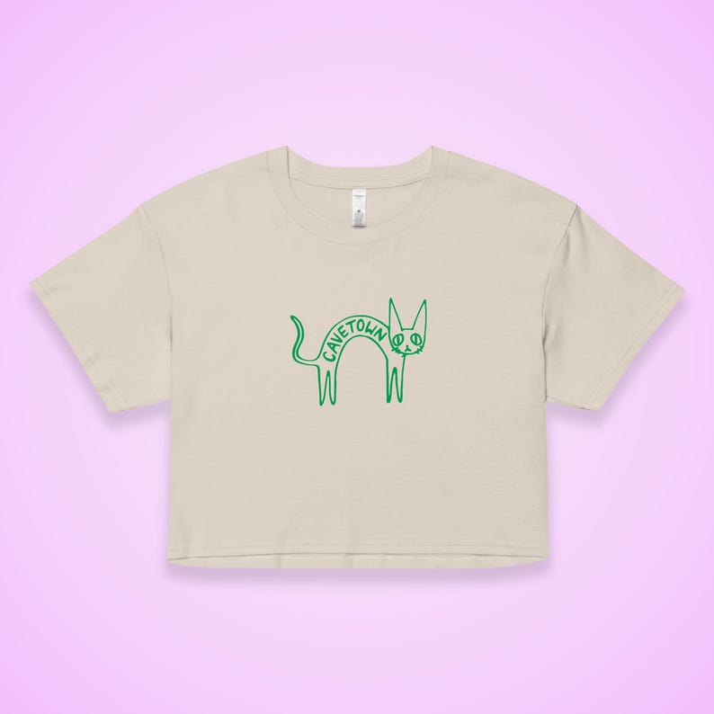 May include: A beige crop top with a green outline drawing of a cat. The word "CAVETOWN" is arched over the cat's back. The cat has a long tail and is standing on its hind legs. The shirt has short sleeves and a crew neck.
