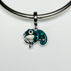 May include: A teal and white chameleon charm suspended from a silver-coloured necklace. The charm features black eyes and rests on a small silver branch. The chameleon's body is a glossy teal colour.