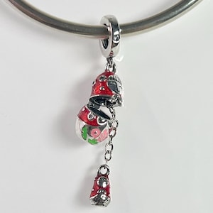 Könnte beinhalten: Ein silberner Charm mit einem rot-weißen Emaille-Design, das eine schneemannartige Figur mit einem durchsichtigen Glaskörper zeigt, der mit rosa und grünen floralen Akzenten gefüllt ist. Ein kleinerer, passender Charm hängt darunter.