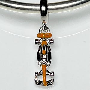 Peut inclure: Un charm argenté en forme de voiture de course de Formule 1 avec des accents orange et noirs. Le charm est suspendu à une boucle argentée, conçu pour un bracelet ou un collier.