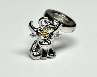 May include: A silver charm featuring a Highland cow with gold-colored details on its head. The charm has a loop for attaching to a bracelet or necklace. The cow is in a sitting position, with detailed horns and fur.