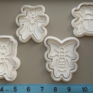 May include: Four white plastic insect-shaped cookie cutters, including a beetle, ant, fly, and butterfly. The cutters are on a grey surface with a ruler at the bottom. The ruler shows measurements in centimetres.