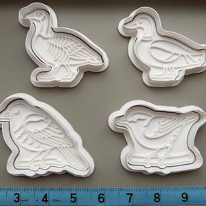 May include: Four white plastic bird-shaped cookie cutters. The cutters include a goose, a duck, and two other bird designs. A blue and white measuring tape is at the bottom of the image.