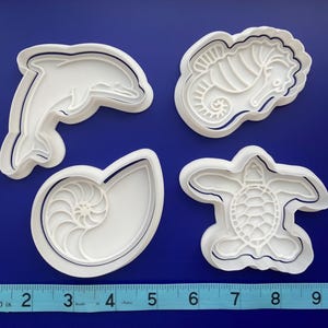 May include: Set of four white plastic cookie cutters shaped like a dolphin, seahorse, nautilus shell, and sea turtle. The cutters are on a blue background with a ruler for scale.