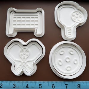 May include: Four white plastic cookie cutters in the shapes of pickleball equipment. The shapes include a net, a paddle, a paddle with a ball, and a ball. The cookie cutters are on a brown surface, with a ruler below.