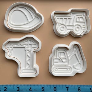 May include: Four white plastic cookie cutters in the shapes of construction vehicles and equipment. The set includes a hard hat, dump truck, crane, and excavator. The cutters are on a tan surface.