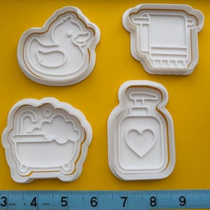 May include: Four white plastic cookie cutters in the shapes of a rubber duck, a towel, a bathtub with bubbles, and a soap dispenser with a heart. The cutters are on a yellow background. A ruler is at the bottom.