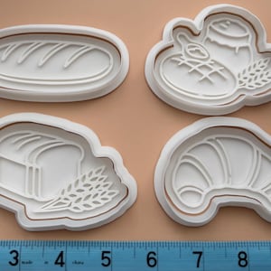 May include: Four white plastic cookie cutters in the shapes of bread and baked goods. The cutters include a loaf of bread, a croissant, a loaf of sliced bread with wheat, and a bread roll with a jar of honey and wheat.
