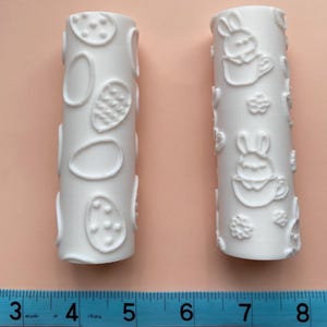 May include: Two white rolling pins with raised designs. One has a pattern of eggs and the other has a pattern of bunnies.