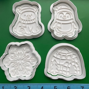 May include: Four white plastic cookie cutters in winter-themed shapes: a bear wearing a scarf, a penguin with a hat and scarf, a snowflake, and an igloo with a Christmas tree. A ruler shows the cutters are about 3 inches wide.