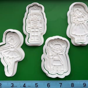 May include: Four white plastic cookie cutters in the shapes of characters from The Nutcracker ballet. The shapes include a ballerina, a nutcracker soldier, a princess, and a mouse king. The cookie cutters are on a green surface. The ruler shows measurements in inches.