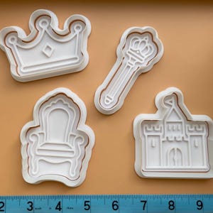 May include: Four white plastic cookie cutters in the shapes of a crown, scepter, throne, and castle. The cutters have a brown outline and are arranged on a tan surface. A ruler is at the bottom of the image.