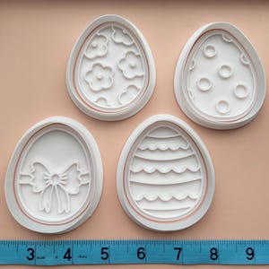 May include: Set of four white plastic Easter egg cookie cutters. The cutters feature different designs, including a bow, polka dots, stripes, and flowers.