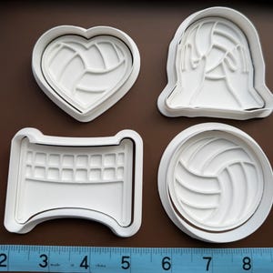 May include: Four white plastic cookie cutters in various shapes related to volleyball. The shapes include a heart with a volleyball design, a volleyball with hands, a volleyball net, and a round volleyball. A ruler is at the bottom.