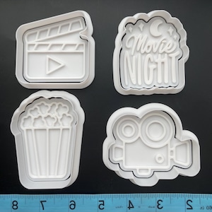 May include: Four white plastic cookie cutters shaped like a movie clapboard, a popcorn bucket, a film camera, and a sign that says "Movie Night".