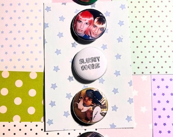 May include: Five circular pinback buttons arranged vertically on a white card with blue stars. The buttons feature a person with dark hair, two people with pink and blonde hair, the text SLUSHY NOOBZ, a person in a hat, and two people in a car.