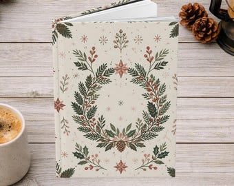 May include: A hardcover journal with a cream-colored cover featuring a central botanical wreath illustration. The design includes green pine branches, red berries, and small star-shaped accents. The book is slightly open, revealing white pages.