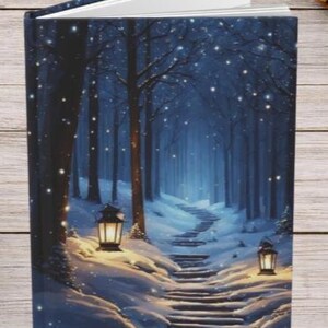 May include: A hardcover book featuring a dark blue winter forest scene on the cover, depicting a snow-covered stone staircase winding through trees, illuminated by two glowing lanterns, with white snow falling against the deep blue night sky.