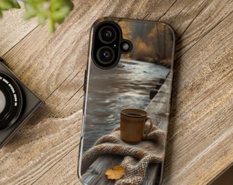 May include: A smartphone case featuring a printed image of a brown ceramic mug resting on a knitted beige scarf atop a wooden dock, with a river and autumn trees in the background. The case has a black camera cutout for a dual lens system.