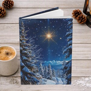 May include: A hardcover book featuring a dark blue night sky illustration with a bright glowing star, snow-covered pine trees, and a winter landscape. The book is slightly open, revealing white pages, and is positioned upright on a wooden surface.