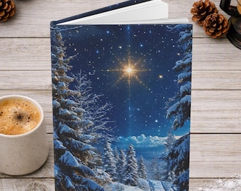 May include: A hardcover book featuring a dark blue night sky illustration with a bright glowing star, snow-covered pine trees, and a winter landscape. The book is slightly open, revealing white pages, and is positioned upright on a wooden surface.