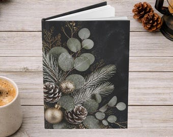 May include: A hardcover journal featuring a dark black cover with a printed design of green eucalyptus leaves, pine branches, brown pinecones, and a gold-toned spherical ornament. The book is positioned upright with white pages visible.