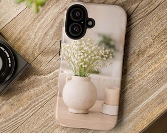 May include: A phone case featuring a printed image of a round, cream-colored ceramic vase filled with white baby's breath flowers, positioned next to a small cylindrical candle on a light-colored surface, with a black camera lens cutout.