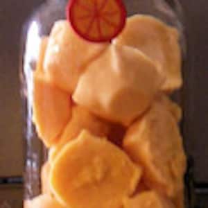 Op de afbeelding: Een glazen pot gevuld met oranje zelfgemaakte zeepjes. De pot heeft een wit deksel en een klein oranje etiket met een afbeelding van een sinaasappelschijfje.