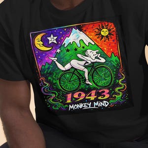 Op de afbeelding: Zwart t-shirt met een kleurrijke afbeelding van een persoon op een fiets voor een berg, zon en maan. De tekst "1943 Monkey Mind" staat eronder, in een psychedelische stijl.