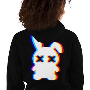 Può includere: Una felpa nera con cappuccio che presenta una grafica bianca di un coniglio sul retro. Il design del coniglio ha un effetto glitch con contorni rossi, blu e gialli e due forme a X scure per gli occhi, creando un aspetto di aberrazione cromatica.