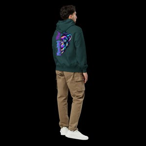 Könnte beinhalten: Ein dunkelgrüner Kapuzenpullover mit einer leuchtenden, neonfarbenen Grafik eines Affengesichts auf der Rückseite, begleitet von dem vertikalen Schriftzug "Monkey" in einer stilisierten violetten Schrift. Dazu eine beige Cargohose und weiße Sneaker.