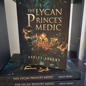 May include: A stack of three paperback books titled The Lycan Prince's Medic by Ashley Argent, with one copy standing upright. The cover features a gold crown surrounded by dark green foliage and glowing orange light effects against a black background.