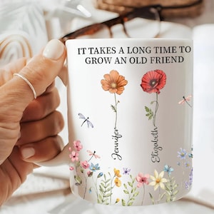 May include: White ceramic mug with the text "IT TAKES A LONG TIME TO GROW AN OLD FRIEND". The mug features floral designs, including orange and red flowers, dragonflies, and the names "Jennifer" and "Elizabeth".