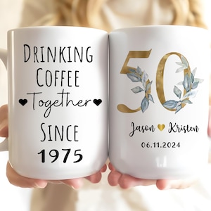 A person is holding two white mugs with text and designs on them.