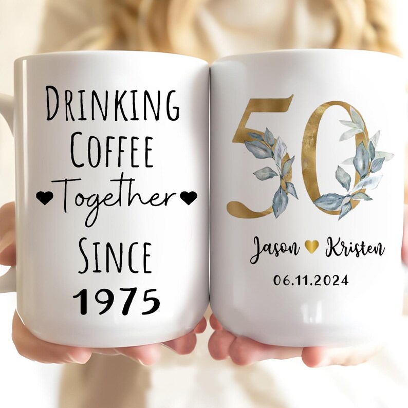 A person is holding two white mugs with text and designs on them.