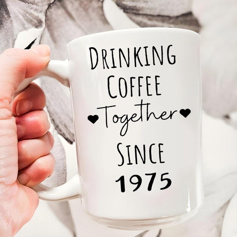 A person's hand holding a white mug with black text that reads "Drinking Coffee Together Since 1975".