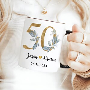 A person is holding a white mug with a gold and blue floral design, and the mug has the number 50 and the names 'JASON' and 'KRISTEN' printed on it.