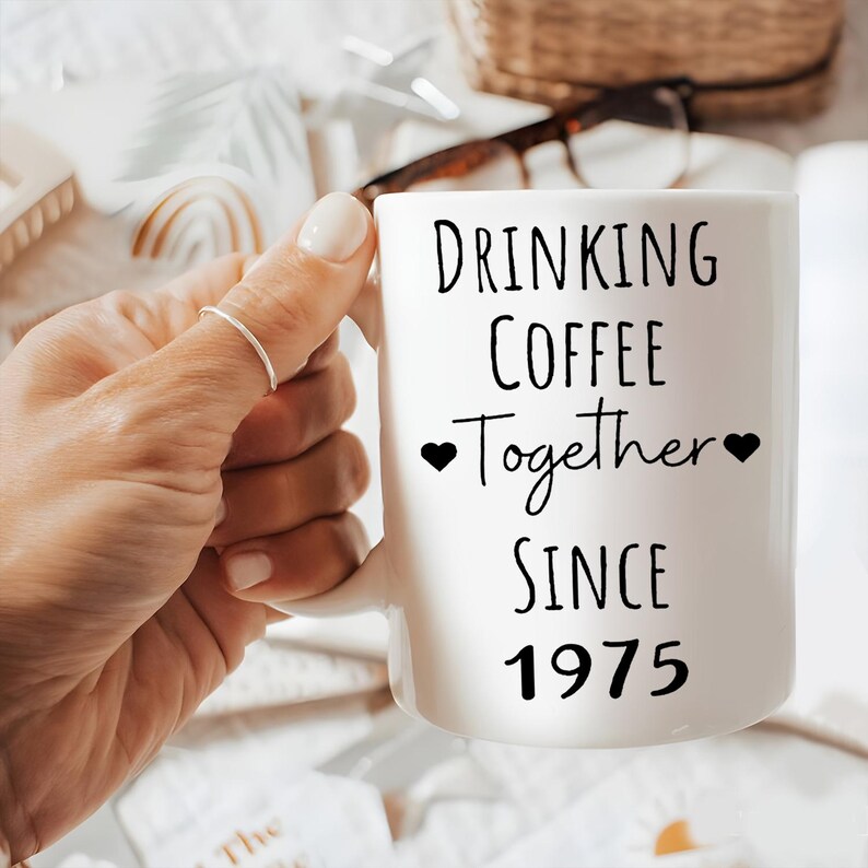 A person's hand holding a white mug with the text "Drinking Coffee Together Since 1975" printed on it.
