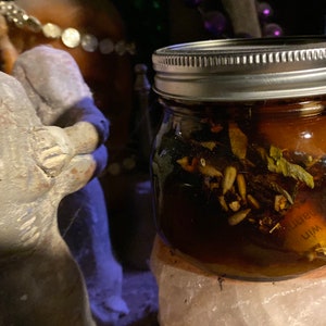 May include: A small glass jar with a silver lid filled with a dark liquid and herbs. The jar sits on a pink crystal. A statue is in the background.