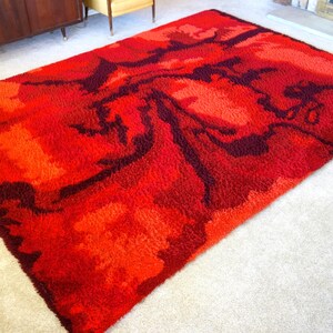 May include: A red and burgundy shag rug with a swirling abstract pattern. The rug is made of a thick, plush material and has a modern, mid-century design.